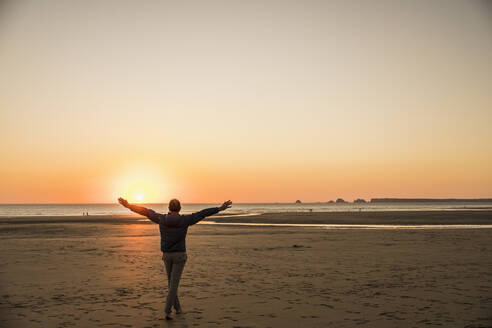 Man with arms outstretched enjoying on shore at sunset - UUF27278