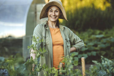 Smiling woman with hand on hip wearing hat in garden - IEF00079