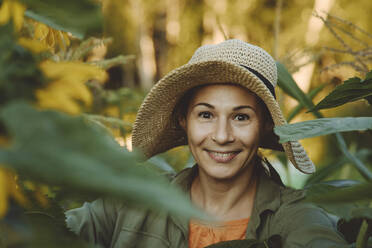 Lächelnde Frau mit Hut inmitten von grünen Blättern im Garten - IEF00086