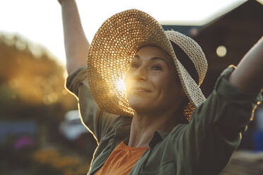 Smiling woman with arms raised wearing hat in garden - IEF00094