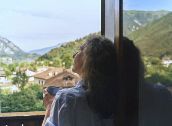 Nachdenkliche Frau mit Kaffeetasse, die am Fenster lehnt - DIKF00695