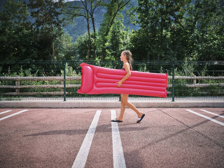 Smiling girl walking with inflatable pink pool raft in parking lot - DIKF00700
