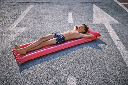 Shirtless boy lying on inflatable pool raft in parking lot - DIKF00703