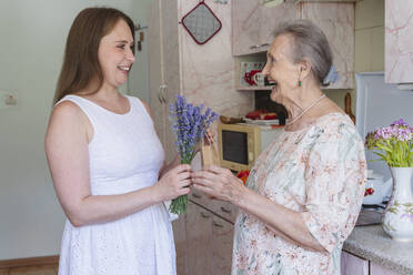 Smiling woman giving bunch of lavender flowers and gift to grandmother at home - OSF00948