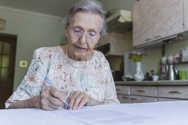 Senior woman with eyeglasses signing home insurance papers on table - OSF00958