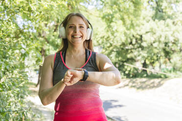 Smiling woman with wireless headphones checking pulse on fitness tracker in park - OSF00999
