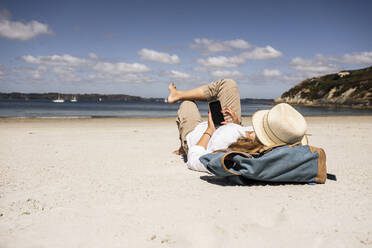 Frau mit Rucksack und Smartphone am Strand liegend - UUF27288