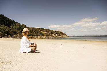 Ältere Frau mit Hut praktiziert Meditation am Strand an einem sonnigen Tag - UUF27292