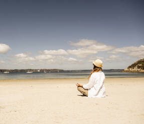 Frau mit Hut praktiziert Meditation am Strand an einem sonnigen Tag - UUF27293
