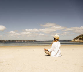 Woman wearing hat practicing meditation at beach on sunny day - UUF27293