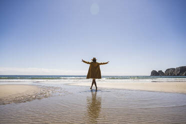 Woman with arms outstretched walking in water at beach on sunny day - UUF27306