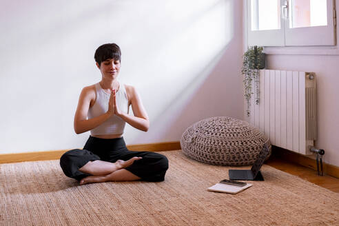 Frau sitzt mit geschlossenen Augen und übt Yoga mit einem Tablet-PC zu Hause - MMPF00290