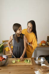 Mother looking at daughter cutting vegetables on board at home - LESF00267