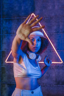 Frau mit drahtlosen Kopfhörern macht Stop-Geste vor Dreiecksform Neonlicht an der Wand - YTF00101