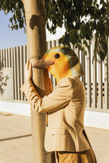 Freiberufler mit Vogelmaske, der an einem sonnigen Tag einen Baum am Fußweg umarmt - MGRF00783