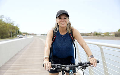 Glückliche ältere Frau mit Mütze auf dem Fahrrad auf der Promenade - FLLF00709