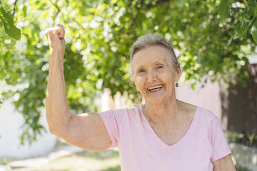 Happy senior woman flexing muscles in front of tree at park - OSF01011
