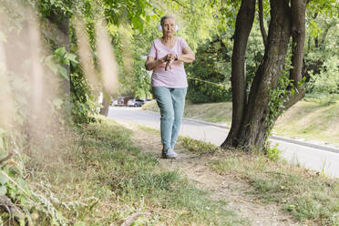 Aktive ältere Frau mit drahtlosen Kopfhörern, die an einem Baum im Park spazieren geht - OSF01029