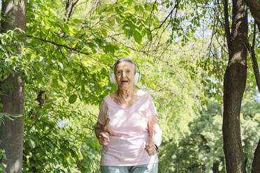 Happy senior woman with wireless headphones jogging amidst trees - OSF01030