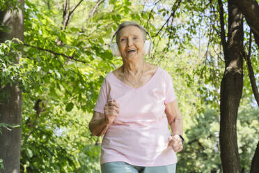 Happy active senior woman with wireless headphones jogging amidst trees - OSF01031