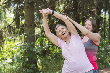 Fröhliche Fitnesstrainerin, die eine ältere Frau beim Training vor einem Baum im Park anleitet - OSF01044