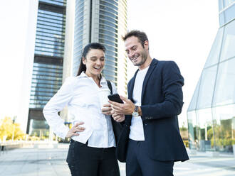 Happy businessman with businesswoman using smart phone standing in front of office building - AMRF00056