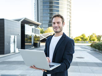 Smiling businessman with laptop standing at office park - AMRF00062