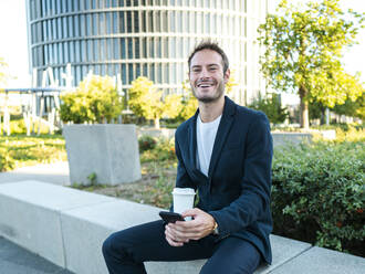 Glücklicher Geschäftsmann mit Einweg-Kaffeebecher und Smartphone im Büropark sitzend - AMRF00064
