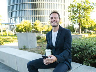 Happy businessman with disposable coffee cup and smart phone sitting at office park - AMRF00064