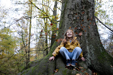 Fröhliches Mädchen sitzt auf einem Baumstamm und beobachtet den Laubfall im Wald - FLLF00724