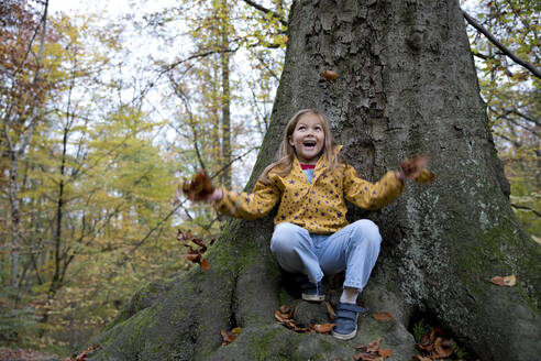 Surprised girl playing with leaves sitting on tree trunk in forest - FLLF00725