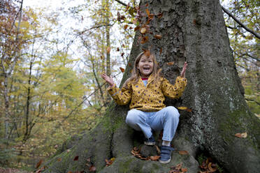 Fröhliches Mädchen spielt mit Blättern auf einem Baumstamm im Wald sitzend - FLLF00726