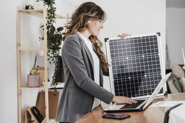 Modern businesswoman working on laptop using portable solar module in office - EBBF06523