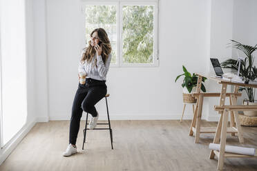 Young businesswoman using mobile phone in modern office holding reusable coffee cup - EBBF06568