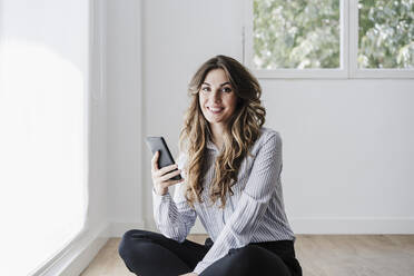 Lächelnde Geschäftsfrau im Büro, die ein Mobiltelefon am Fenster benutzt - EBBF06569