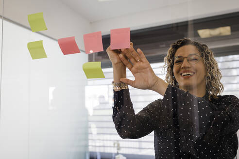 Happy mature businesswoman writing on sticky notes on glass at office - DCRF01490
