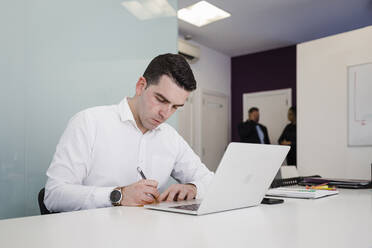 Young businessman writing in front of laptop at desk in office - DCRF01495