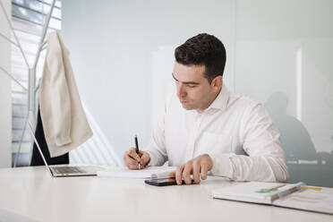 Junger Geschäftsmann mit Schrift in Buch sitzend am Schreibtisch im Büro - DCRF01496