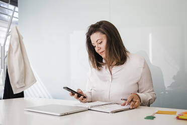 Reife Geschäftsfrau mit Smartphone am Schreibtisch im Büro - DCRF01499