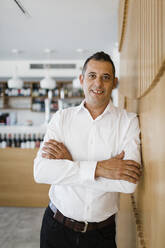 Smiling businessman with arms crossed leaning on wall at restaurant - DCRF01533