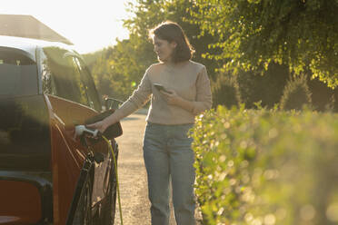 Woman with mobile phone charging electric car at roadside - EKGF00157