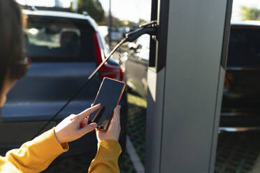 Woman using smart phone at electric vehicle charging station on sunny day - EKGF00160