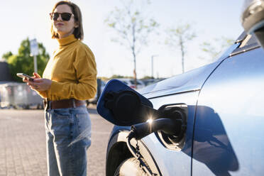 Electric car charging at station with woman standing in background on sunny day - EKGF00171