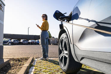 Elektroauto an einer Ladestation mit einer Frau im Hintergrund, die an einem sonnigen Tag ihr Smartphone hält - EKGF00173