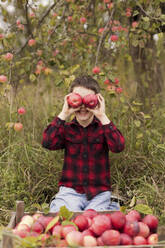 Smiling boy holding red apples over eyes by crate - ONAF00140