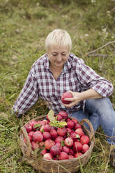 Reife Frau mit Korb voller Äpfel im Gras sitzend auf einem Bauernhof - ONAF00148