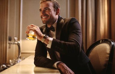 Lächelnder junger Mann bei einem Drink in einem Nachtclub. Mann im formellen Anzug in einem Nachtclub, der ein Glas Whiskey an der Bar trinkt. - JLPPF00004