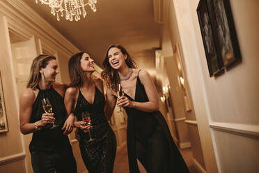 Eine Gruppe von drei Frauen mit Champagnergläsern geht durch einen Korridor, beste Freundinnen auf einer Gala. - JLPPF00058
