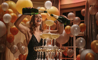 Fröhliche junge Frau füllt Champagner in einen Glasturm auf einer Gala. Aufgeregte Frau im Abendkleid füllt eine Pyramide von Gläsern mit Champagner. - JLPPF00081
