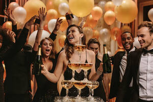 Pyramide von Champagner mit Menschen tanzen um in der Partei. Gläser in Turmform mit Champagner und Gruppe von verschiedenen Freunden tanzen im Hintergrund auf Neujahrsparty gefüllt angeordnet. - JLPPF00083